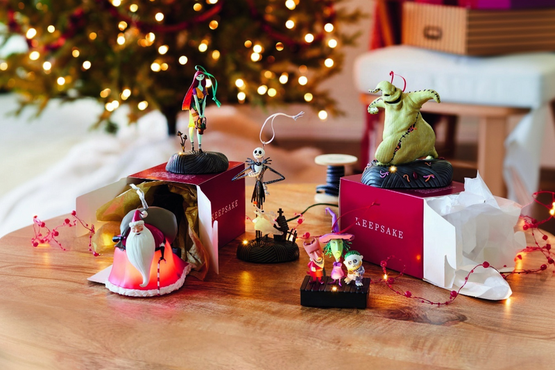 Hallmark Keepsake ornaments of Sally, Jack Skellington, and Oogie Boogie on a table with a glowing tree background and red gift boxes.