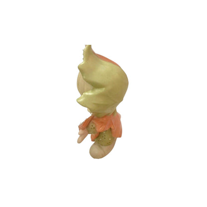 Small figurine of a character with a green head and peach-colored outfit on a white background